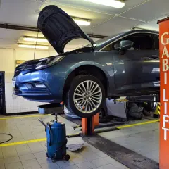 W warsztacie samochodowym trwa wymiana oleju i filtrów. Auto uniesione na podnośniku, pod nim widać miskę olejową.