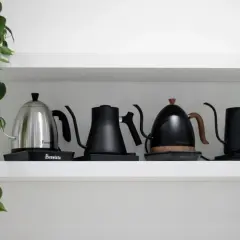 Fyra vattenkokare, en i metall och tre i svart, står på en hylla. En grön växt hänger ner från vänster. Perfekt för att koka te vatten.