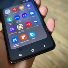 Dłoń trzyma smartfon z widocznym folderem aplikacji, w tym ikoną Bixby.