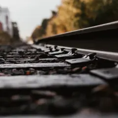 Zbliżenie na podkłady kolejowe i śruby mocujące szyny. W tle widać zamazane budynki i drzewa, sugerujące drogę przebiegu.