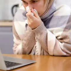 Kobieta w kratę, z chusteczką w dłoni, korzysta z laptopa. Może to być polskie zwolnienie lekarskie w Danii, gdy czuje się źle.