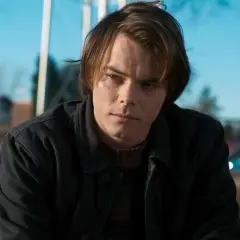 Charlie Heaton w ciemnej kurtce i swetrze, zamyślony, na tle błękitnego nieba i samochodów.