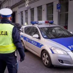 Policjant w kamizelce odblaskowej stoi obok radiowozu z napisem "POLICJA".