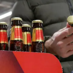 Rząd butelek piwa w czerwono-złotych etykietach. Człowiek w zielonej kurtce trzyma jedną butelkę, zastanawiając się, ile kosztuje jedno piwo.