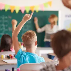 Uczniowie z podniesionymi rękami słuchają uważnie, co mówi wychowawca przy tablicy.