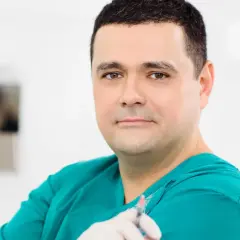 Lekarz w zielonym fartuchu trzyma strzykawkę. W tle widać elementy gabinetu. Wiele pozytywnych klinika ambroziak opinie potwierdza jego profesjonalizm.