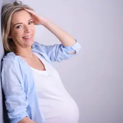 Uśmiechnięta kobieta w ciąży po 40. opiera się o ścianę, dotykając włosów.