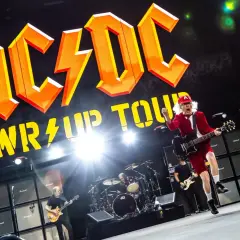 AC/DC na koncercie w Polsce. Wokalista w czerwonym stroju skacze z gitarą na tle gigantycznego logo zespołu i ściany wzmacniaczy.