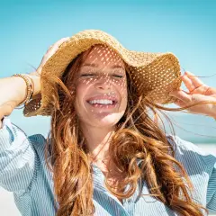 Mujer sonríe bajo sombrero de paja, con el pelo suelto. Consejos para como cuidar el pelo en verano.