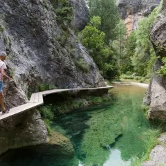 Hombre camina por pasarela de madera en la ruta parrizal de beceite, junto a un río de aguas cristalinas y paredes rocosas.