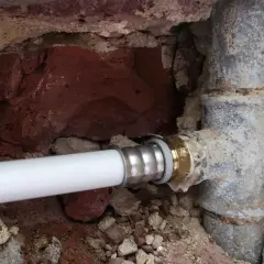 Un nouveau tuyau blanc est raccordé à un ancien tuyau de plomb pour l'eau. La maçonnerie est visible autour des canalisations.