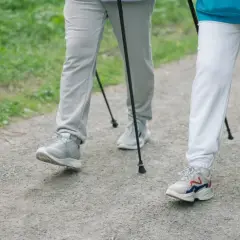 Dwie osoby uprawiające **nordic walking** na żwirowej ścieżce, otoczone zielenią.