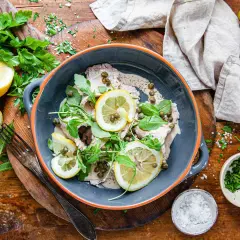 Vitello tonnato i en grå skål, garnerad med citronskivor, kapris och ruccola. Serveras med färska örter och citroner på ett träbord.