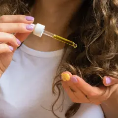 Eine Hand mit lila und gelbem Nagellack hält eine Pipette mit goldenem Ricinus Communis Seed Oil über lockiges Haar.
