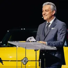 Hombre sonriente en traje, presentando un coche amarillo eléctrico, quizás un nuevo modelo de Volvo Renault.