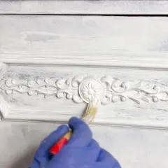 Mano con guanto blu che applica chalk paint fai da te su un mobile con decorazioni in rilievo, creando un effetto shabby chic.