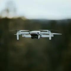 Biały dron DJI Mini 2 w locie, z widocznym obiektywem kamery. Tło to rozmyty las i niebo.