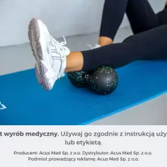 Kobieta wykonuje rolowanie mięśni łydki za pomocą czarnego rollera z zielonymi kropkami, leżąc na niebieskiej macie.