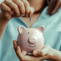 Ręka wrzuca monetę do różowej świnki-skarbonki.