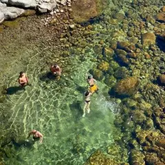 Gente se baña en un río cristalino con fondo de piedras, cerca de un **ponton de la oliva**.
