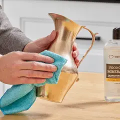 Hands polish a brass pitcher with a blue cloth, showing how to clean brass with mineral oil.