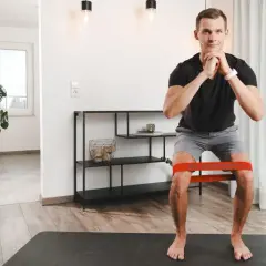Mann macht Kniebeugen mit Widerstandsband. Die tiefe Flexion im Knie wird durch das Band unterstützt und erschwert.