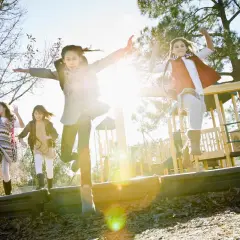 Vier fröhliche Kinder springen ausgelassen auf einem Spielplatz. Sie lernen in Bewegung, die Sonne scheint, und ihre Arme sind in die Luft gestreckt.