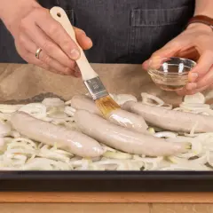 Bratwurst im Backofen: Zwiebelringe auf Backblech, darauf vier rohe Bratwürste. Mit Pinsel wird Öl aufgetragen.