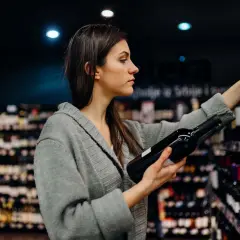 Donna sceglie una bottiglia di vino, pensando ai solfiti. Scaffali pieni di bottiglie sullo sfondo.