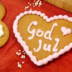 Hjärtformade pepparkakor med rosa glasyr och texten "God Jul". Bredvid syns en pepparkaka dekorerad med vita pepparkakor-liknande mönster.