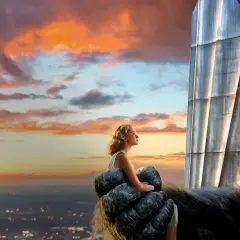 King Kong trzyma kobietę na dłoni, patrząc na zachód słońca nad miastem.