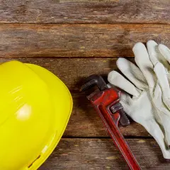 Żółty kask, klucz hydrauliczny i rękawice na drewnianym tle. Zastanawiasz się, dlaczego woda schodzi, a jest bulgotanie w rurach? Czas na fachowca!