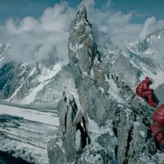 Dwóch alpinistów w czerwonych kurtkach odpoczywa na skalistej grani, podziwiając lodowiec i ośnieżone szczyty. Widok na berbeka broad peak zapiera dech w piersiach.