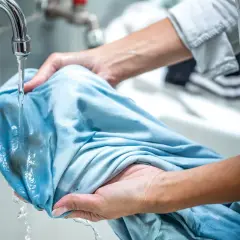 Ręce płuczą niebieską koszulkę pod bieżącą wodą, próbując usunąć plamy. Dowiedz się, czym zmyć farbę do włosów z ubrań.