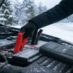Rękawiczka podłącza czerwony kabel do akumulatora. Obok leży urządzenie rozruchowe, które pomoże odpalić samochód w zimowy dzień.