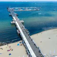 Słoneczny dzień nad Bałtykiem. Molo w Polsce tętni życiem, spacerują po nim tłumy ludzi. Na plaży odpoczywają plażowicze, a w oddali widać jachty.