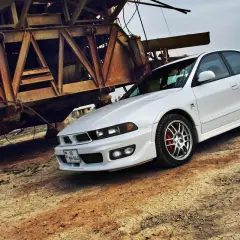 Biały Mitsubishi Galant, lśniący obok zardzewiałej, industrialnej konstrukcji, prezentuje się elegancko na tle suchej ziemi.