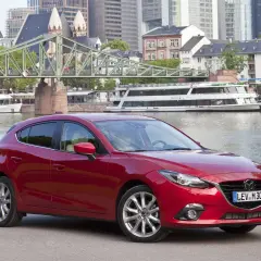 Czerwona Mazda 3 na tle miasta i rzeki. Wiele osób szuka takich aut, bo mazda 3 opinie ma zazwyczaj bardzo dobre.