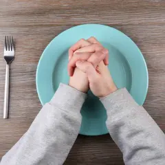 Manos entrelazadas sobre plato vacío, tenedor y cuchillo a los lados. Quizás piensa que no cenar adelgaza.