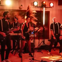 Zespół gra na scenie, wokalistka z burzą włosów, gitarzysta i klawiszowiec. W tle biały miś tekst.
