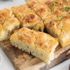 Focaccia dorada y tierna con sal gruesa y romero, cortada en porciones sobre tabla de madera.