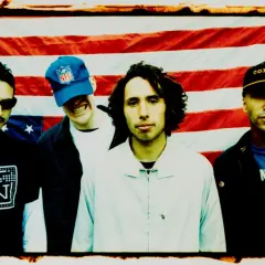 Członkowie Rage Against the Machine pozują przed amerykańską flagą. Jeden z nich ma na głowie czapkę z napisem "commie".