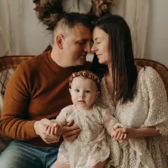 Ciepła sesja na roczek z rodzicami. Malutka córeczka w koronkowej sukience trzyma dłonie mamy i taty, którzy patrzą na nią z miłością.