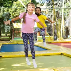 Dwie dziewczynki skaczą na trampolinie, śmiejąc się radośnie. Czy trampolina jest zdrowa dla dziewczynek? Z pewnością tak, to świetna zabawa i aktywność fizyczna!