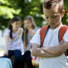 Ein Junge mit Rucksack steht traurig da, während zwei Mädchen ihn im Hintergrund auslachen. Ein trauriges Bild von Mobbing in der Grundschule.