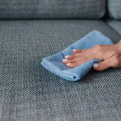 A hand with white nail polish wipes a grey couch with a blue microfiber cloth, demonstrating how to remove slime from couch.