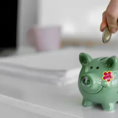Ręka wrzuca monetę do zielonej świnki skarbonki. To symbol oszczędzania i finansowego zabezpieczenia.