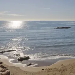 Costa de Villaricos con un faro en un rompeolas de rocas. El sol brilla sobre el mar azul y la arena dorada.