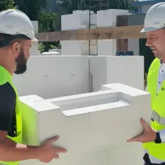 Dwóch budowlańców w kaskach i kamizelkach przenosi element konstrukcyjny. Kierownik budowy może wprowadzić zmiany w projekcie, np. dotyczące materiałów.