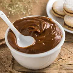 Un tazón blanco lleno de dulce de leche cremoso, con una cuchara blanca. Al fondo, alfajores espolvoreados con azúcar glas.